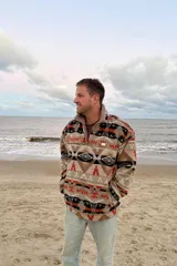 Buzo de polar con diseño estampado de motivos aztecas en tonos beige, naranja, negro y blanco. Presenta cuello alto con cierre frontal parcial y corte holgado.