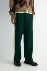 Pantalón deportivo de corte relajado en tejido efecto neopreno, color verde oscuro con franjas laterales en contraste color crema. Presenta cintura elástica con cordón ajustable, bolsillos laterales y traseros, y cierre oculto en el bajo.