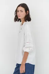 Camisa de color crudo con diseño romántico, cuello redondo con cordón ajustable terminado en borlas, y mangas tres cuartos abullonadas. Presenta detalles de bordados calados verticales en el frente y horizontales en las mangas.