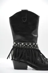 Bota de cuero negro estilo texano, con taco bajo y punta fina. Incluye un aplique desmontable en la zona del empeine decorado con tachas metálicas y flecos largos.