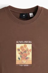 Remera marrón de algodón con cuello redondo, mangas cortas y hombros caídos, de corte holgado. Presenta un estampado frontal con la obra 'Los girasoles' de Van Gogh y el texto 'Sunflowers' en la parte superior y '-Van Gogh-' en la inferior.