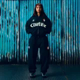 Campera con capucha y cierre frontal, color negro, con estampado gráfico frontal que incluye el logo de la marca y un escudo estilo deportivo.