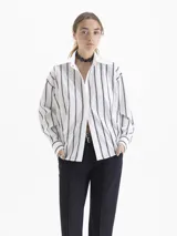 Camisa blanca de poplin con fit holgado, cuello con broche metálico y mangas largas con puños abotonados. Presenta un patrón de rayas verticales formadas por bordados de boquitas.