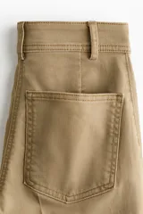 Pantalón de sarga color tostado, de tiro alto y corte acampanado con diseño tobillero. Presenta bolsillos frontales tipo parche con solapa abotonada y pliegues marcados al frente y en la espalda.