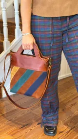 Bolso tipo matera confeccionado en cuero vacuno, con diseño estructurado y manija superior integrada. Presenta un cuerpo con franjas diagonales en gamuza de colores contrastantes (amarillo, verde y azul) sobre una base de cuero marrón, con detalles de costuras artesanales en los bordes.