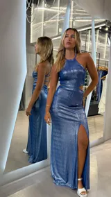 Vestido largo de color azul con acabado metalizado, cuello halter y diseño con aberturas (cut-out) en la cintura. Presenta una falda larga con tajo lateral profundo y espalda descubierta con tiras finas cruzadas.