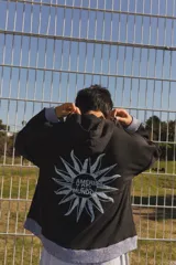 Campera negra de algodón con capucha, cierre frontal con doble cursor y bolsillos laterales. Presenta puños y ruedo tejidos en color celeste. En la espalda, destaca un estampado de un sol con la frase 'De América y del mundo'.