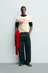 Camiseta de corte regular, color crema, con cuello redondo y ribetes en las mangas y cuello en color negro. Presenta un estampado frontal con un corazón rojo y texto en negro que dice "Everything beats for something".