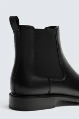 Bota estilo Chelsea de cuero negro con diseño liso, paneles elásticos laterales y tirador trasero. Presenta horma redonda y suela con tacón bajo.