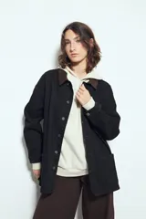 Sobrecamisa tipo Barn Jacket color negro, de algodón con elastano, con silueta relajada y estructura firme. Presenta cierre frontal con botones metálicos, cuello de pana marrón, bolsillos aplicados y pespuntes visibles en color marrón.