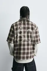 Sobrecamisa de corte relajado confeccionada en algodón, con estampado de cuadros en tonos marrones y beige. Presenta cuello solapa, manga corta, cierre frontal de botones y un bolsillo plastrón en el pecho.