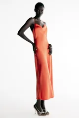 Vestido midi satinado de color naranja tostado, con escote fluido tipo cowl y tirantes finos cruzados en la espalda con detalle de aplicación metálica. Presenta un diseño largo con tajo lateral en la falda.