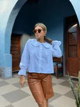 Camisa blanca de corte holgado con cuello bebé con volados, cierre frontal de botones y mangas largas con puños terminados en volados.