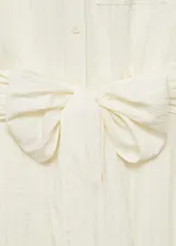 Vestido midi de corte evasé, sin mangas y con cuello camisero. Presenta cierre frontal con botones, un bolsillo de parche en el pecho y un lazo decorativo en la cintura. Confeccionado en tejido con textura de rayas verticales.