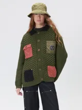 Campera tejido grueso y abrigado color verde militar, con forro de nylon interno, cuello estilo camisa en color negro y cierre frontal con botones. Cuenta con cuatro bolsillos delanteros con apliques en distintos tonos y texturas, y un parche bordado circular en el bolsillo superior izquierdo.