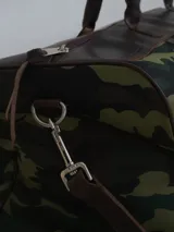 Bolso de viaje tipo duffel de lona negra con detalles en cuero tostado, asas de mano y correa ajustable para el hombro.