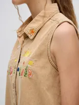 Camisa sin mangas tipo musculosa confeccionada en denim liviano de color tostado. Presenta cuello camisero y cierre frontal con botones. Destaca por sus bordados coloridos en el pecho con motivos veraniegos como sol, olas y elementos marinos.