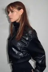 Cazadora de estilo bomber en efecto piel color negro, con cuello subido, manga larga y bajo elástico. Presenta trabillas en los hombros, bolsillos frontales con solapa y cierre de cremallera con botones a presión ocultos.