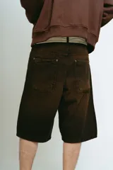 Bermuda de jean color marrón oscuro con efecto lavado, corte baggy y largo hasta la rodilla. Presenta bordados ornamentales en color crema/dorado en los bajos de ambas piernas.