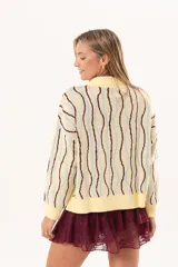 Buzo de punto tipo polo en tono amarillo claro con diseño de ondas a rayas en tonos marrón y celeste. Presenta cuello clásico, cierre frontal parcial con botones y silueta relajada con mangas largas.