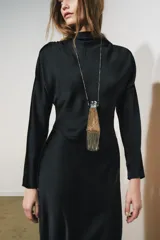 Vestido largo negro de tejido satinado, con cuello subido, manga larga y drapeado en los hombros. Presenta una abertura en la espalda con cierre de botones forrados a tono.