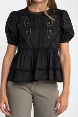 Blusa negra de algodón con corte oversize, mangas cortas abullonadas y detalles de encaje y bordados en el frente y hombros. Presenta un diseño con pliegues verticales y terminaciones con puntilla.