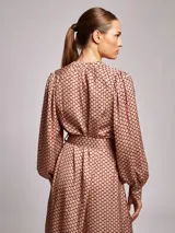 Vestido midi de satén con estampado geométrico en tonos marrones y naranjas. Presenta cuello mao, mangas largas abullonadas con puños ajustados y un cinturón a tono con hebilla metálica que marca la cintura.