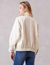 Campera bomber de eco cuero color beige, con cuello camisero, cierre frontal metálico y bolsillos delanteros con solapa y botón a presión. Presenta mangas largas con puños abotonados y ruedo trasero elastizado.