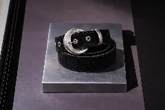 Cinto de cuero negro con textura gamuzada, presenta una hebilla metálica plateada de diseño redondeado con detalles grabados y pasador metálico a juego.