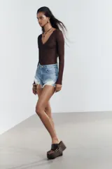 Short de jean negro desgastado, tiro alto, con roturas pronunciadas y terminación deshilachada en el bajo. Presenta cinco bolsillos y cierre frontal con botón metálico.