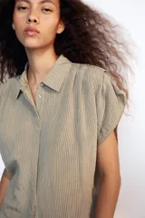 Camisa fluida de cuello solapa y manga corta, con diseño a rayas en tonos beige y blanco, detalle de pliegues en los hombros y cierre frontal con botones.