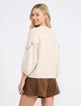 Buzo de punto beige con mangas tres cuartos anchas, cuello redondo y detalle de botones en las mangas.