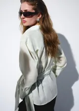 Camisa cruzada de ramio en color crudo, con cuello camisero, escote en V y mangas largas con puños anchos abotonados. Cuenta con un diseño envolvente con detalle de lazo lateral.
