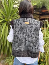 Chaleco reversible con un lado de piel sintética gris y el otro lado de tela bordada con diseño floral y espiral en tonos blanco y negro. Presenta un diseño sin mangas y corte recto.