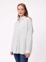 Camisa de corte oversize con diseño a rayas verticales en tonos azul y blanco. Presenta cuello clásico, cierre frontal con botones y un bolsillo en el pecho con detalle de etiqueta.