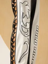 Pantalón de cuero blanco y negro, de corte recto, con detalles de cuero color camel. En los laterales, lleva una pieza de cuero con cordones de cuero negro y camel trenzados. En el delantero lleva bordados de hilo y cuero plata.