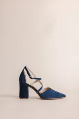 Zapato de tacón color azul marino, con punta fina y escote en V. Presenta tiras cruzadas en el tobillo con hebilla dorada y un taco de 8 cm de altura. Confeccionado en gamuza con forro y plantilla de cuero.