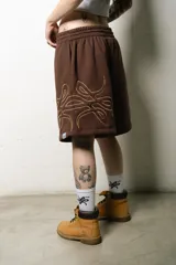 Bermuda estilo baggy de algodón color marrón oscuro, con cintura elástica y cordón blanco. Presenta un gran logo estampado en tono gris claro en la pierna derecha.