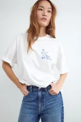 Camiseta de corte oversize, color blanco, con cuello redondo, mangas cortas y estampa botánica pequeña en el pecho.