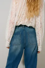 Pantalón de denim azul con lavado medio, corte de pierna recta y tiro alto. Presenta bolsillos de parche delanteros con solapa, trabillas para cinto y detalles de pliegues marcados en el centro de las piernas.