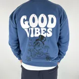 Buzo azul de algodón peletizado, corte oversize, con cuello redondo y mangas largas. Estampa frontal con texto "Good Vibes" y dibujo de un personaje. Estampa trasera con el mismo diseño.