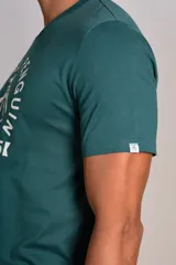 Remera de algodón color verde petróleo con cuello redondo y mangas cortas. Presenta un estampado gráfico frontal en color blanco con el logo de la marca y la leyenda Original Penguin Est. 1955.