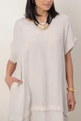 Vestido midi de lino color beige, de corte holgado y silueta acampanada. Cuenta con cuello redondo, mangas cortas amplias, bolsillos laterales y detalles de recortes horizontales en la falda con terminación asimétrica más larga en la parte trasera.