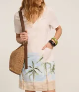 Vestido corto de corte recto y escote en V, color beige claro con estampado de paisaje tropical con palmeras en la parte inferior, mangas cortas y bolsillos laterales. Marca Marfinno.