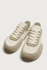 Zapatilla deportiva estilo barefoot, color marrón, con diseño de paneles en combinación de materiales y detalles en serraje. Cuenta con puntera redondeada y ancha, cordones en color crema y suela fina de goma flexible en contraste.