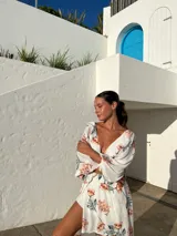 Vestido corto blanco con estampado floral en tonos naranja y verde, escote en V con detalle de nudo, mangas largas acampanadas y falda con volantes y abertura lateral.