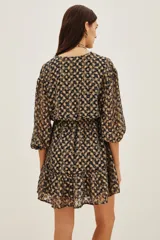 Vestido corto de escote en V cruzado, con mangas tres cuartos y cintura elástica. Presenta un diseño estampado con motivos geométricos en tonos oscuros y detalles metalizados, con un ruedo con volado.
