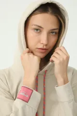 Buzo hoodie color arena, de rústico, con capucha y cordón de ajuste en salmón y verde. Tiene moldería oversize y etiqueta de la marca en la manga derecha con logo Basset 00'S.