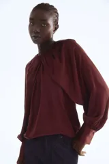 Blusa de satén color borgoña con mangas globo largas y puños anchos. Presenta un detalle de drapeado o nudo en el centro del escote redondo.