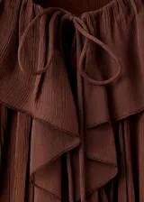 Blusa de color marrón chocolate, confeccionada en tejido plisado con textura. Presenta un diseño de cuello redondo con detalle de lazo, mangas largas amplias con puños acampanados y un corte holgado con volados sutiles en la parte frontal.